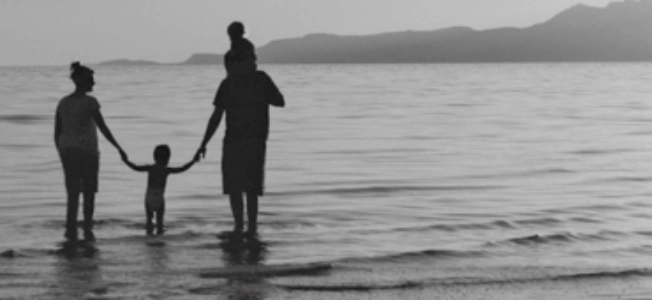 To forældre med mindre børn i vandkant på strand (sort/hvid).